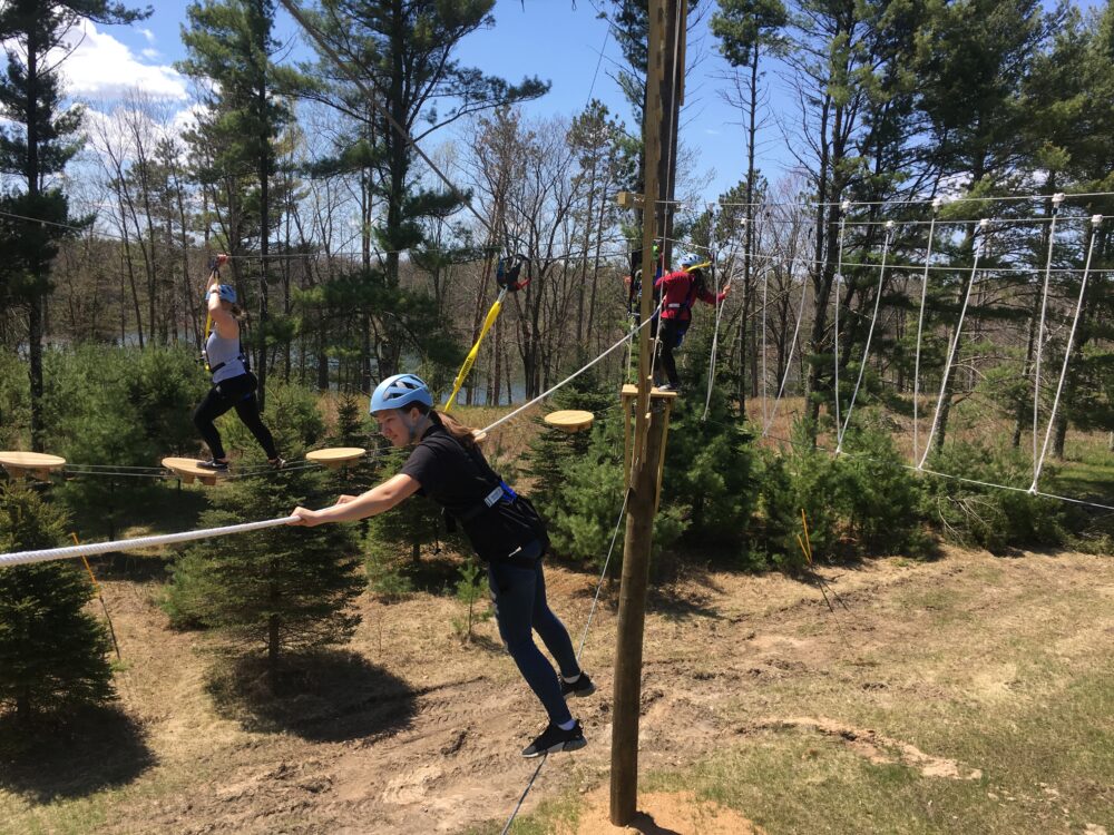 Photo of an individual standing on a wire while holding a rope on the first level of the adventure course