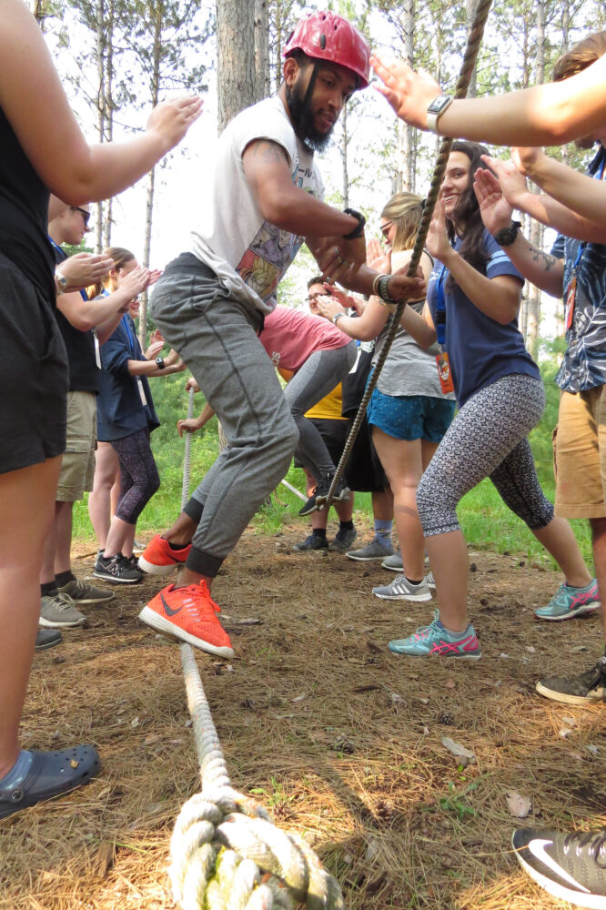 Photo of a person walking on a rope and holding onto a rope while participating in the low ropes course while a group of people are around them spotting them with their hands up