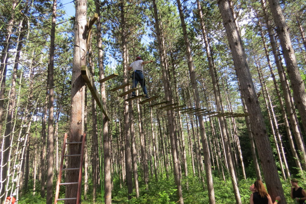A photo of a person walking across the bosun chairs on the shaded high ropes element in the woods